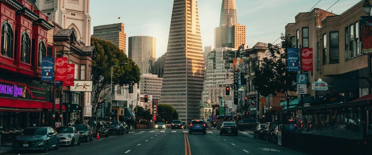 Cars driving down a busy San Francisco street at sunset, with the Transamerica Pyramid and other downtown skyscrapers rising in the background and colorful storefronts lining both sides of the road.