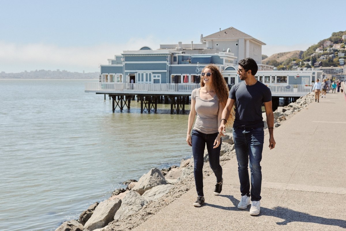 Couple strolling on waterfront path in Tiburon with blue restaurant building on pier and bay views