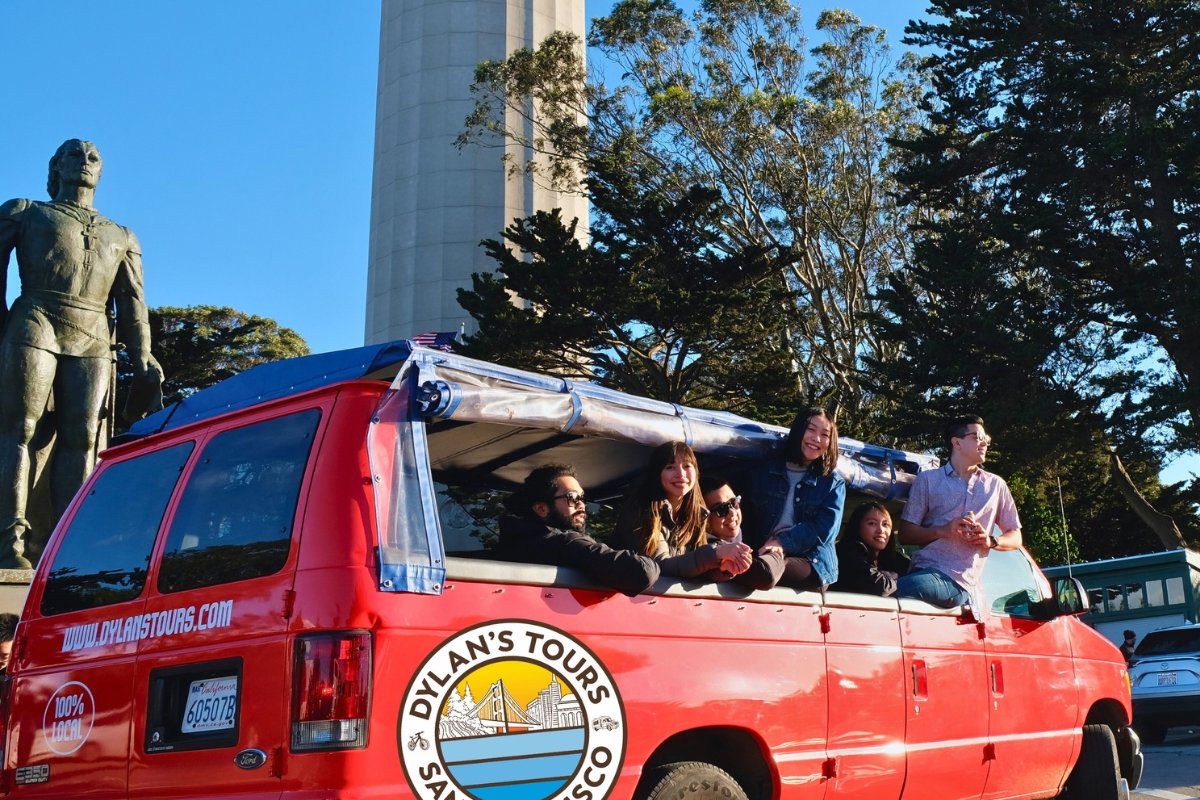 Red Dylan's Tours van with tourists at Coit Tower plaza next to bronze statue overlooking San Francisco skyline