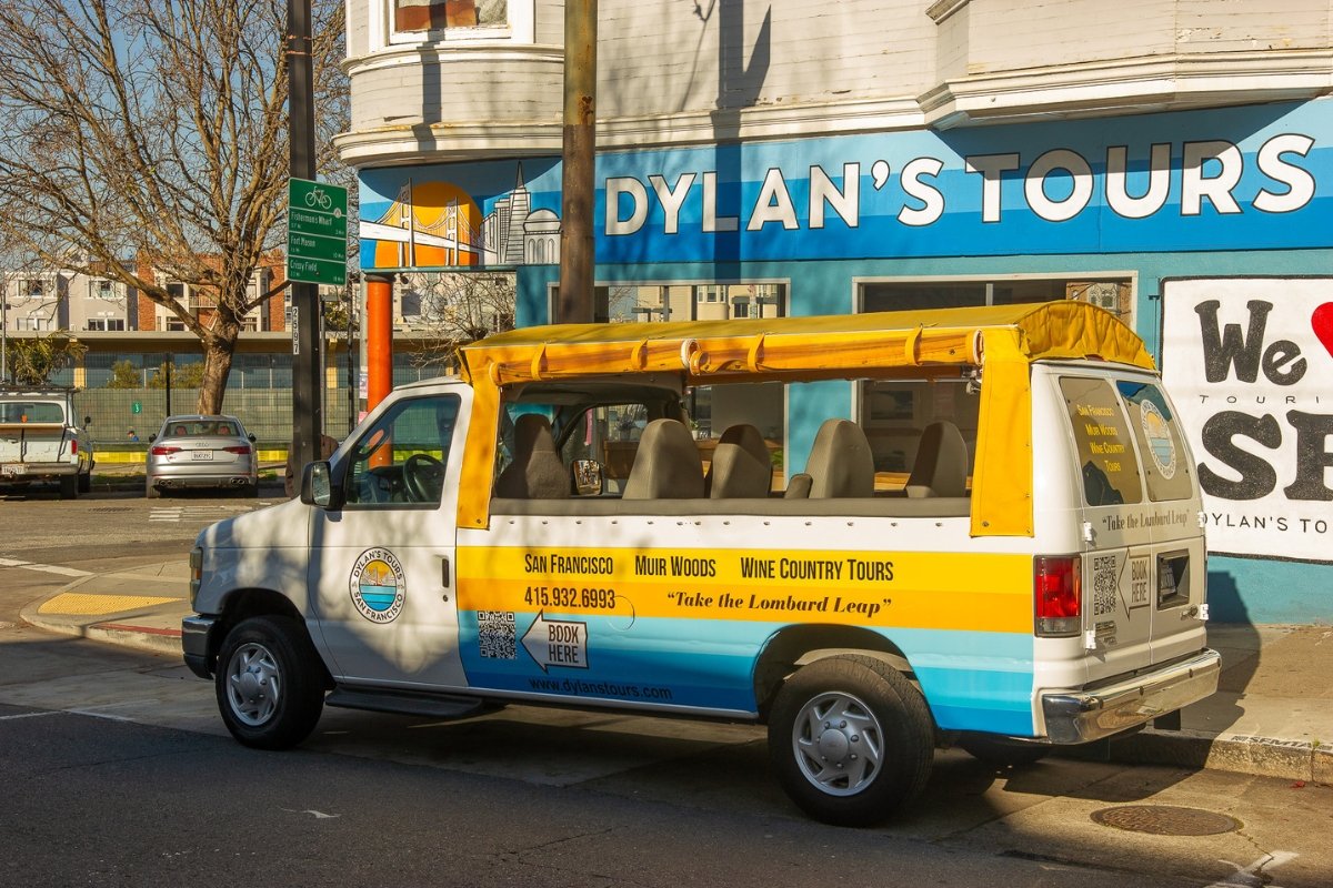 White and blue Dylan's Tours van with yellow roof rack advertising San Francisco, Muir Woods, and Wine Country tours parked outside the company's bright blue storefront.