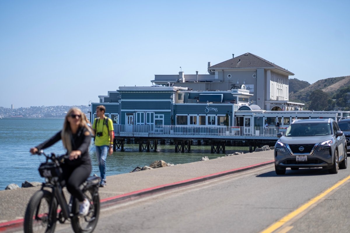 Waterfront restaurant built on pier with cyclist and pedestrian in foreground, blue water and hills in background