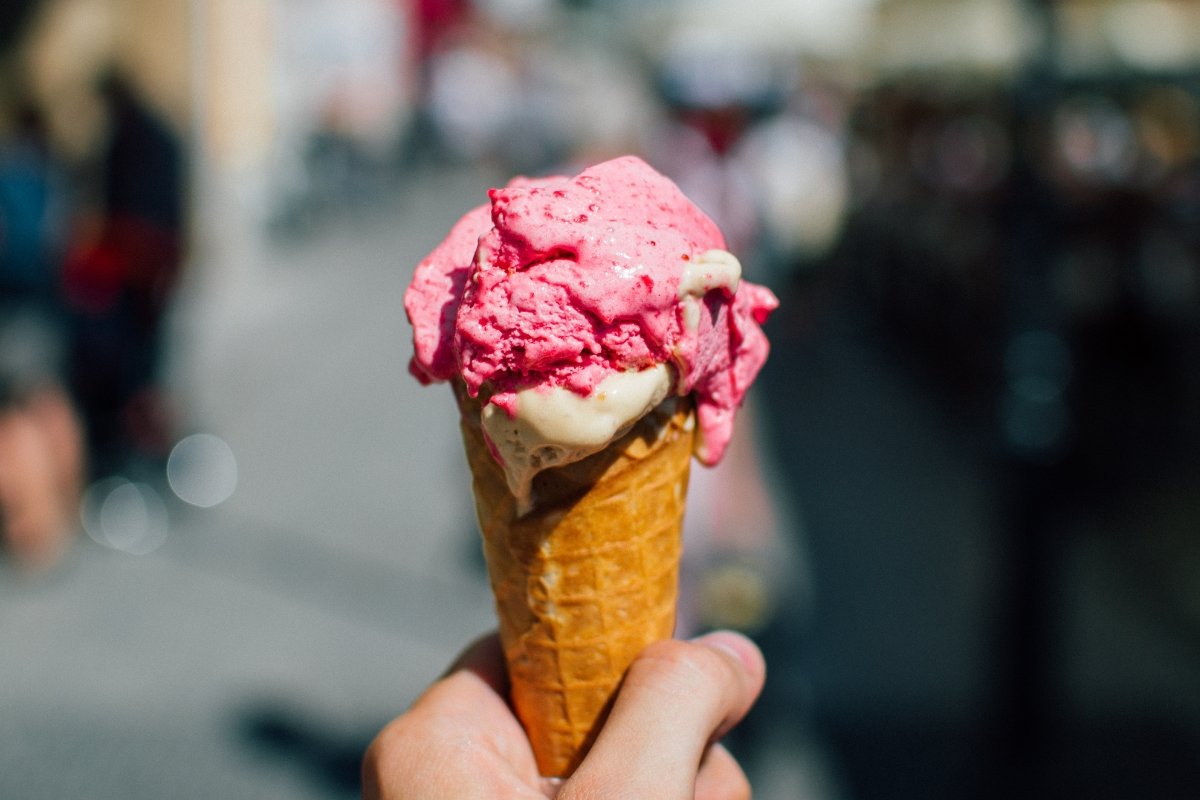 Close-up of a bright pink strawberry or raspberry ice cream scoop with vanilla swirl on a classic waffle cone held by hand