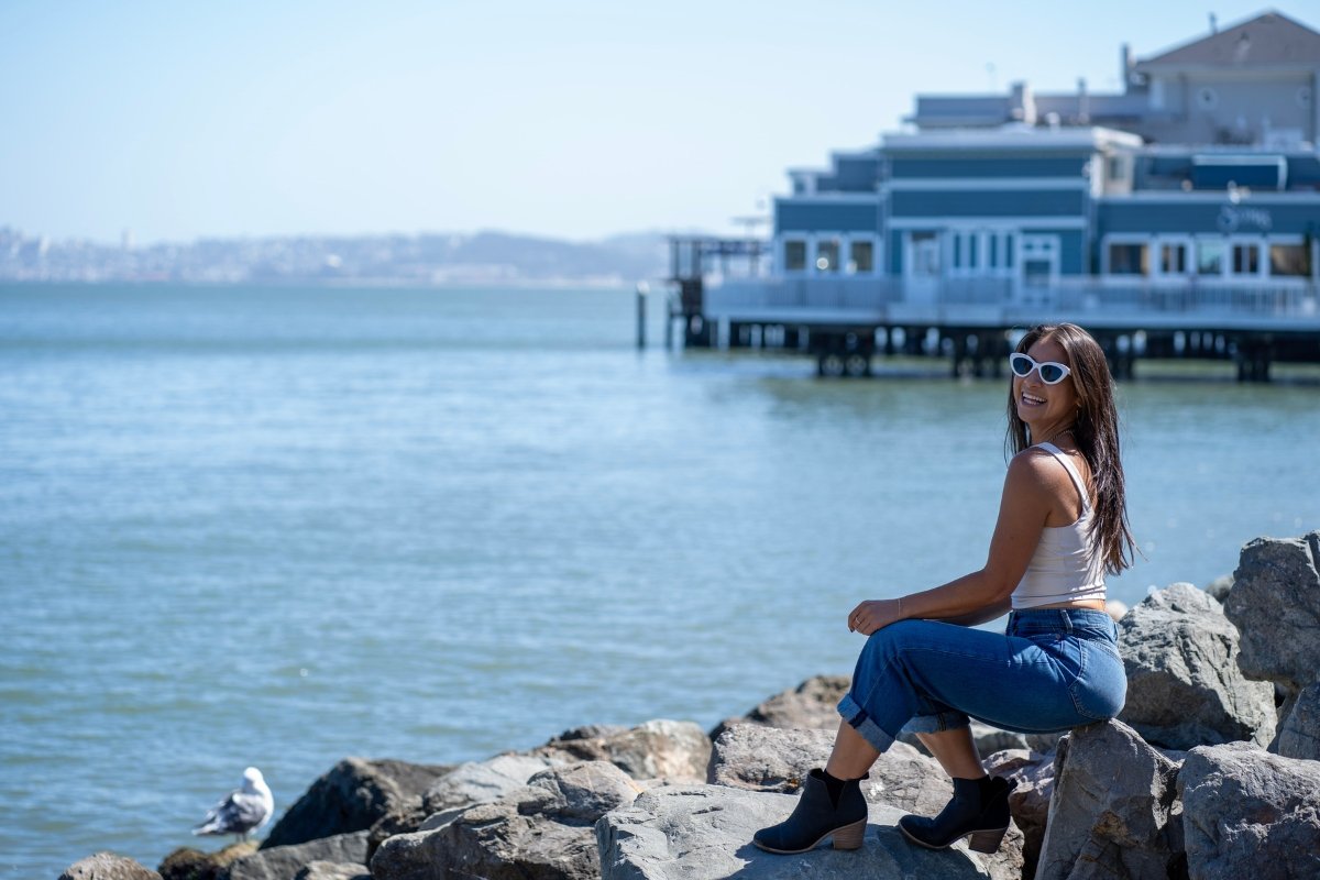 Woman sitting on shoreline rocks in Tiburon with retro sunglasses, waterfront restaurant pier, and Bay waters