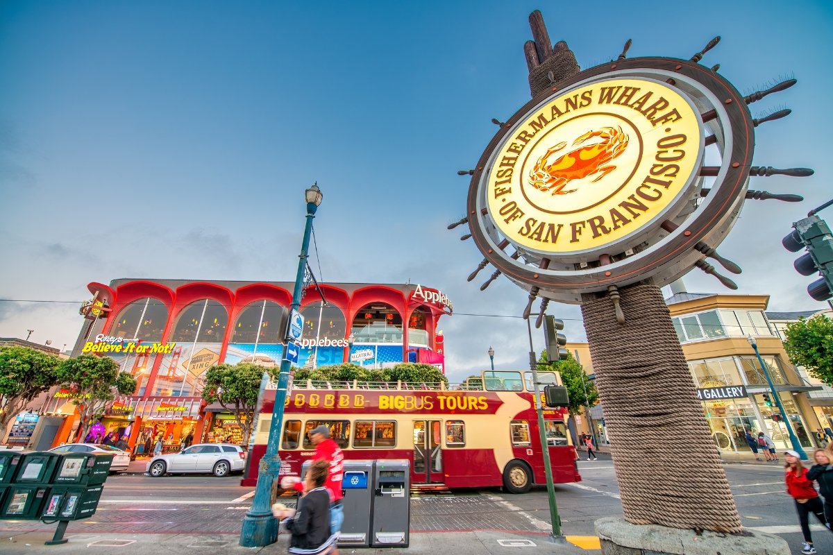 Illuminated Fisherman's Wharf sign with crab logo, Applebee's restaurant, and Big Bus Tours vehicle in San Francisco