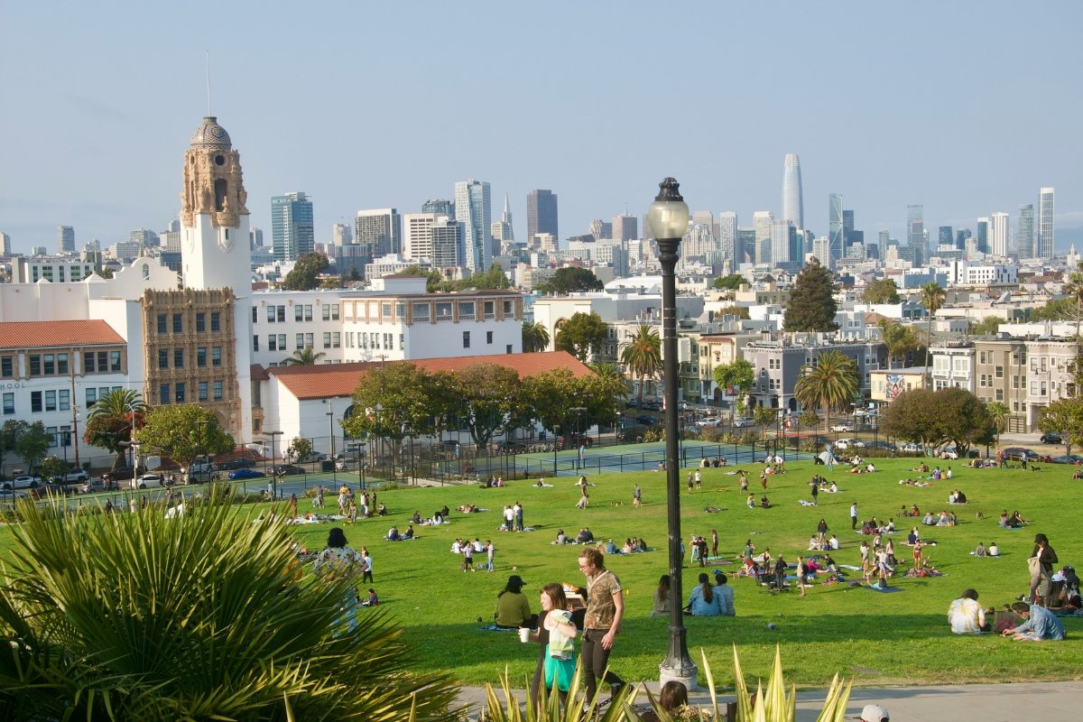 Sunny day at Mission Dolores Park in San Francisco, with people relaxing on green grass, historic Mission-style buildings, and downtown skyscrapers in background