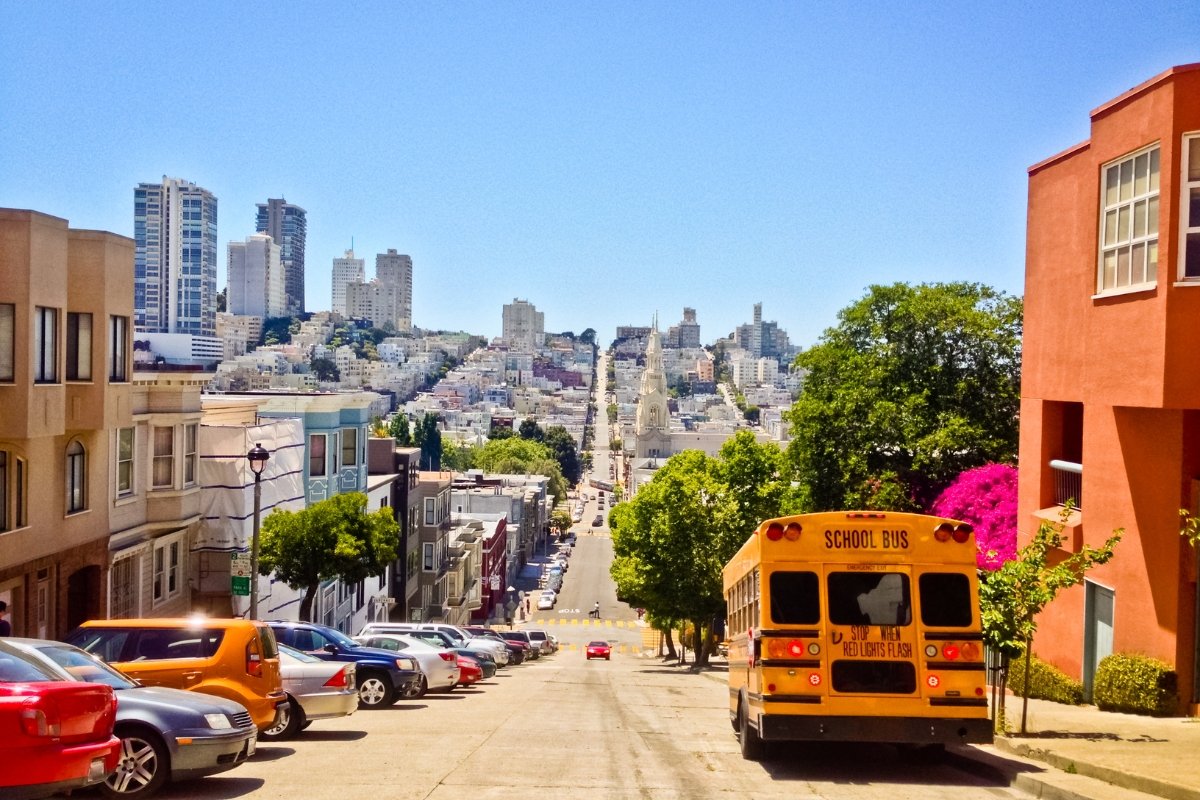 View down a steep San Francisco hill with parked cars, school bus, colorful buildings, and downtown skyline in the distance