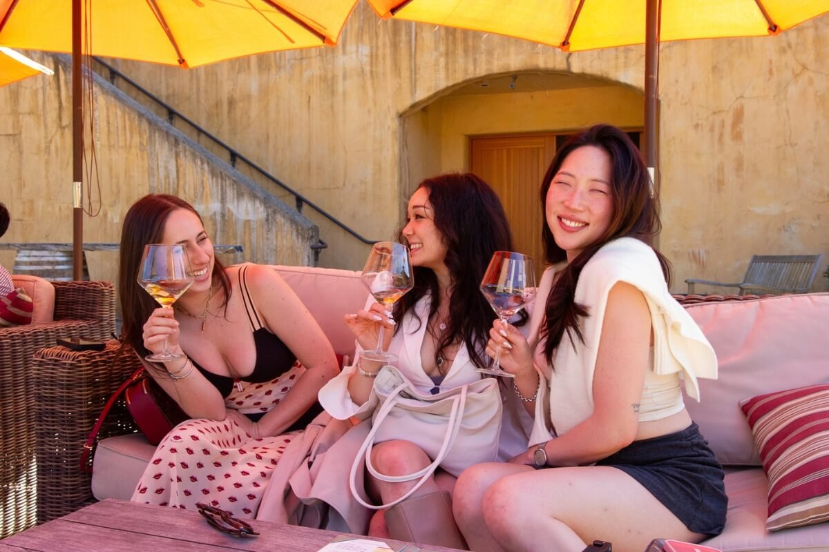 Three women laughing while wine tasting under bright yellow umbrellas on a sunny day at California winery patio.