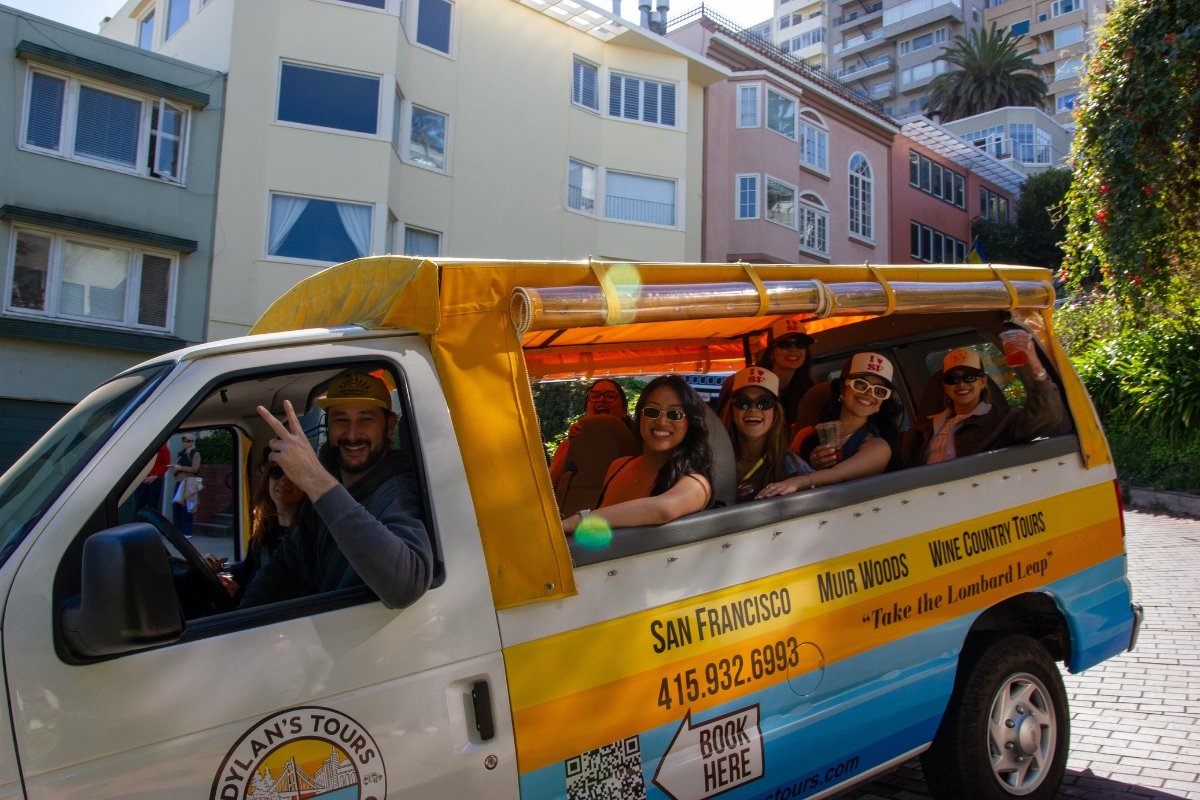 Dylan's Tours van with cheerful tourists wearing San Francisco caps traveling through colorful residential neighborhood with pastel buildings