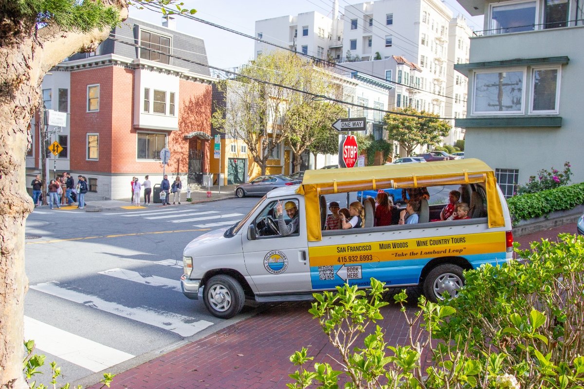Tour van from Dylan's Tours waiting at a San Francisco intersection with stop sign, pedestrians, and classic neighborhood architecture visible