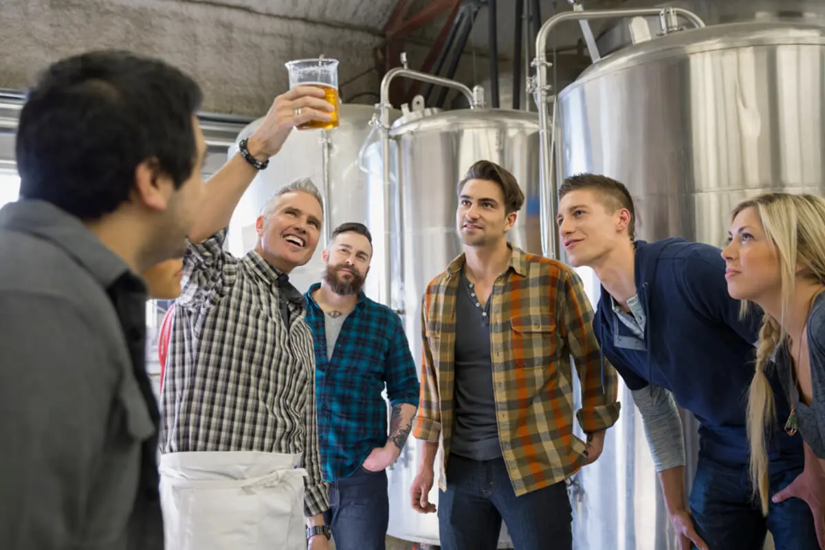 Group of people on a brewery tour listening as a guide holds up a glass of beer for explanation.
