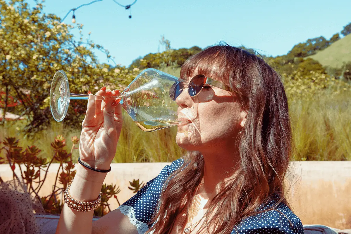 Close-up of a hand holding a glass of white wine outdoors, with sunlight reflecting off the glass and blurred vineyard scenery in the background.