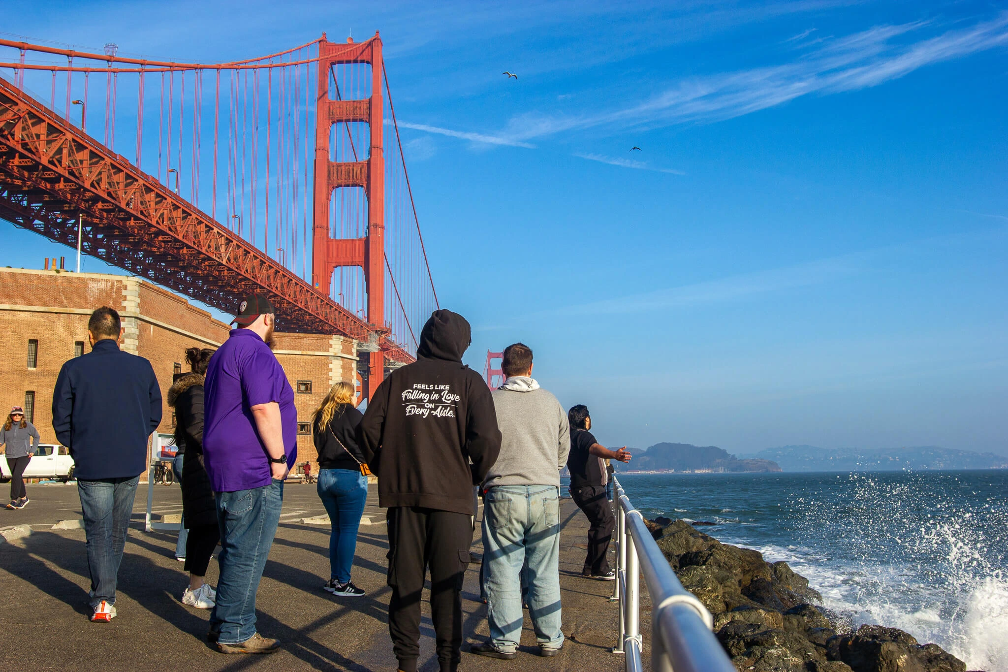 People stroll along the seawall by Fort Point as waves splash against the rocks, with the Golden Gate Bridge towering overhead on a clear blue day.