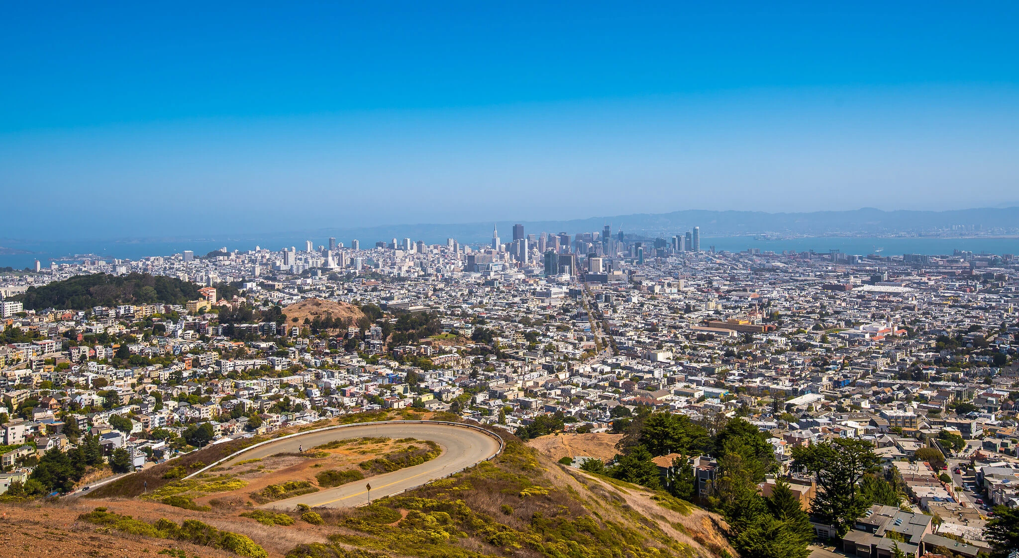 Wide view from Twin Peaks showing a winding road in the foreground, residential neighborhoods below, and downtown San Francisco with the bay in the distance under a clear blue sky.