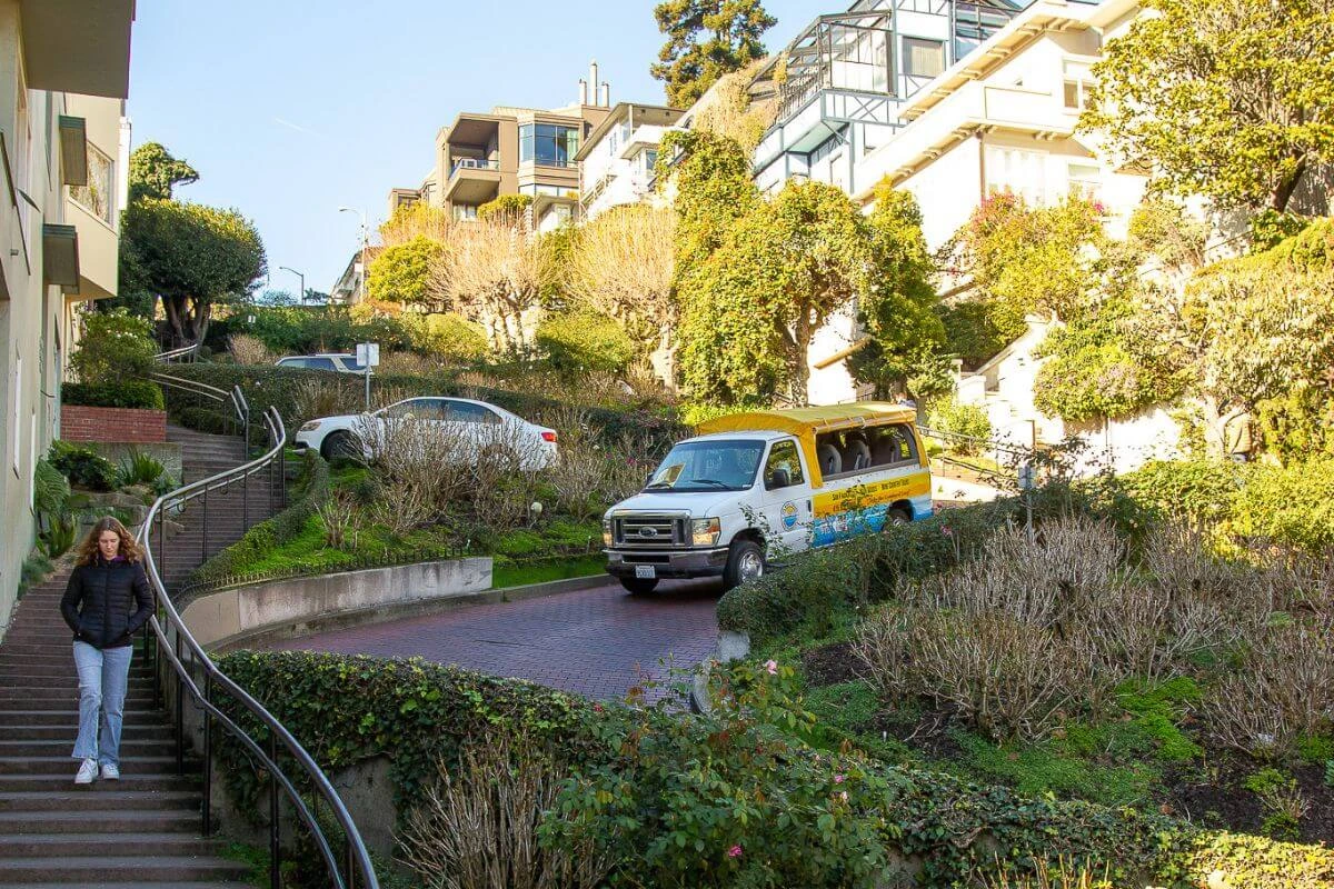 A tour van descends the brick switchbacks of Lombard Street, bordered by landscaped gardens, while a pedestrian walks down the adjacent stairs on a sunny day.
