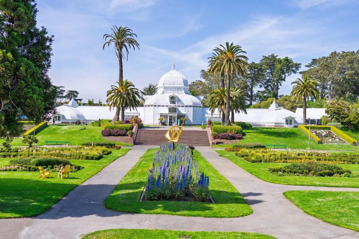 Symmetrical view of the white Victorian glasshouse with palm trees, steps, and landscaped flowerbeds along a central path in Golden Gate Park, San Francisco, on a sunny day.