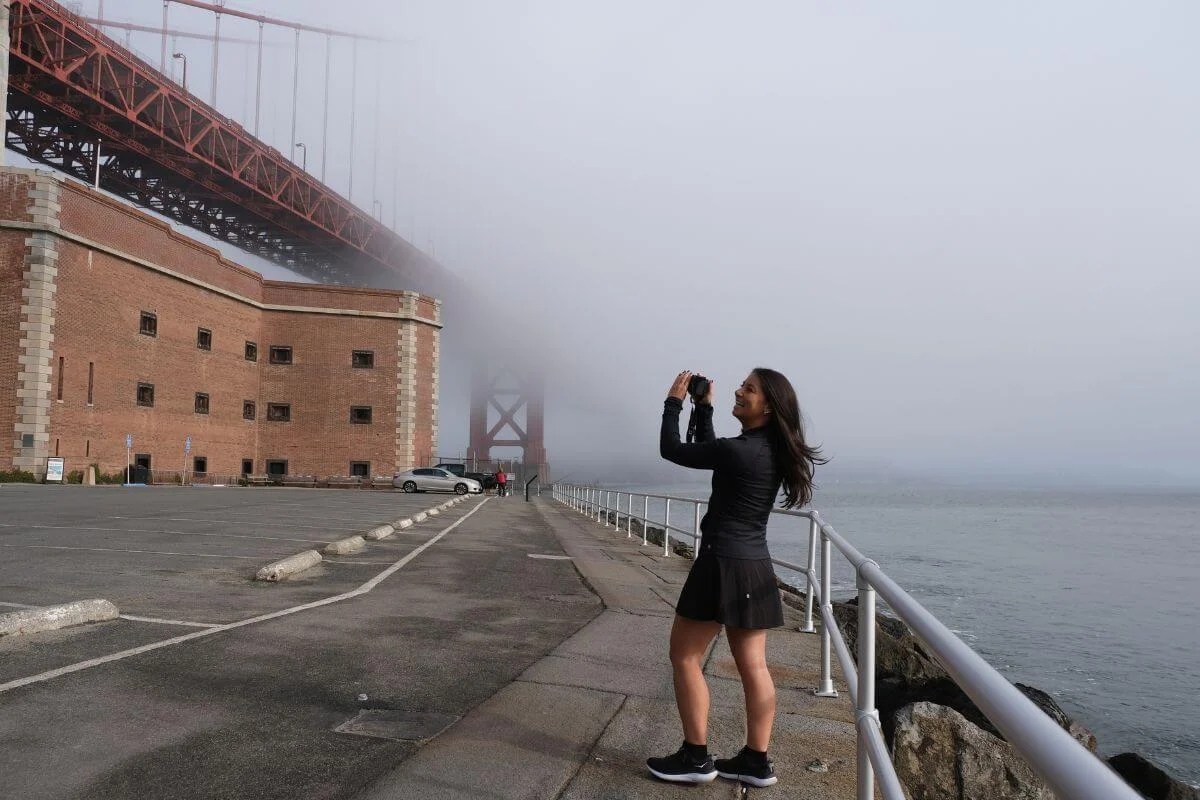 Woman taking a photo on the waterfront path by Fort Point as the Golden Gate Bridge disappears into thick fog over the bay.