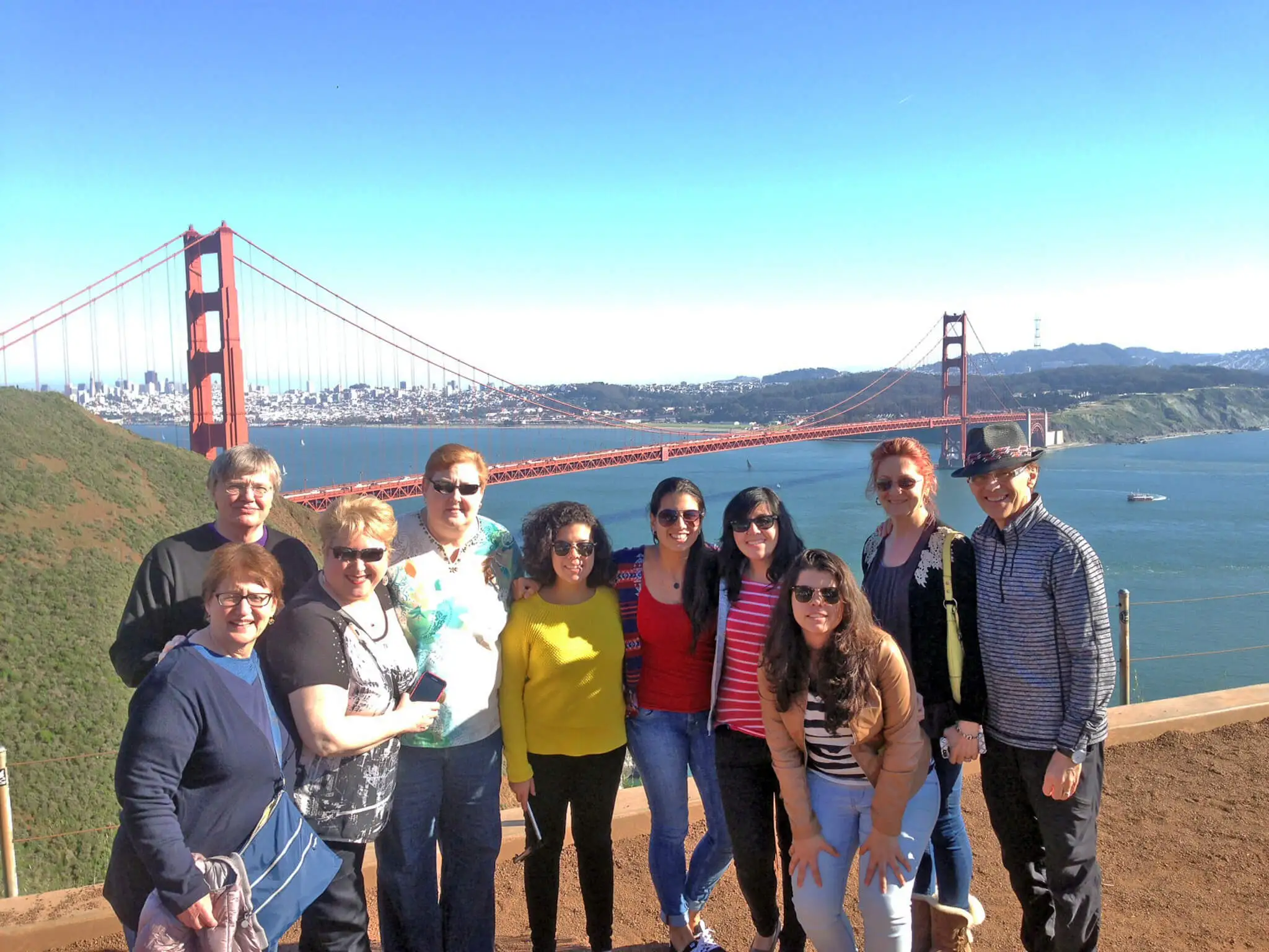 Group of tourists smiling in front of the Golden Gate Bridge on a sunny day with the San Francisco skyline in the background.