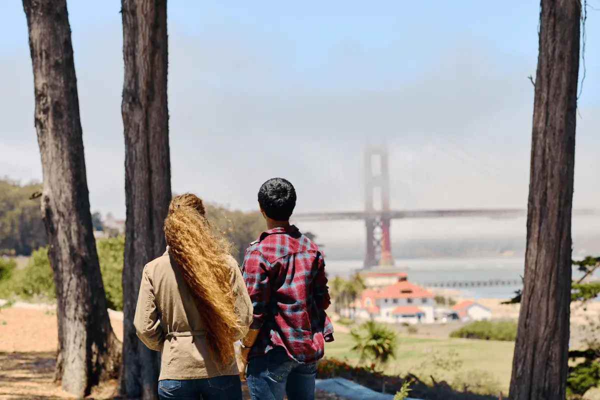 Couple enjoying a scenic view of the Golden Gate Bridge from the San Francisco side, surrounded by trees and coastal fog.