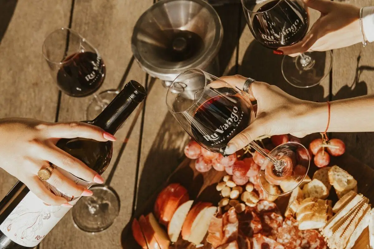 Close-up of hands pouring and holding glasses of red wine labeled “Ru Vango” over a wooden table with a charcuterie board featuring grapes, sliced apples, cured meats, cheese, nuts, and bread under warm natural light.