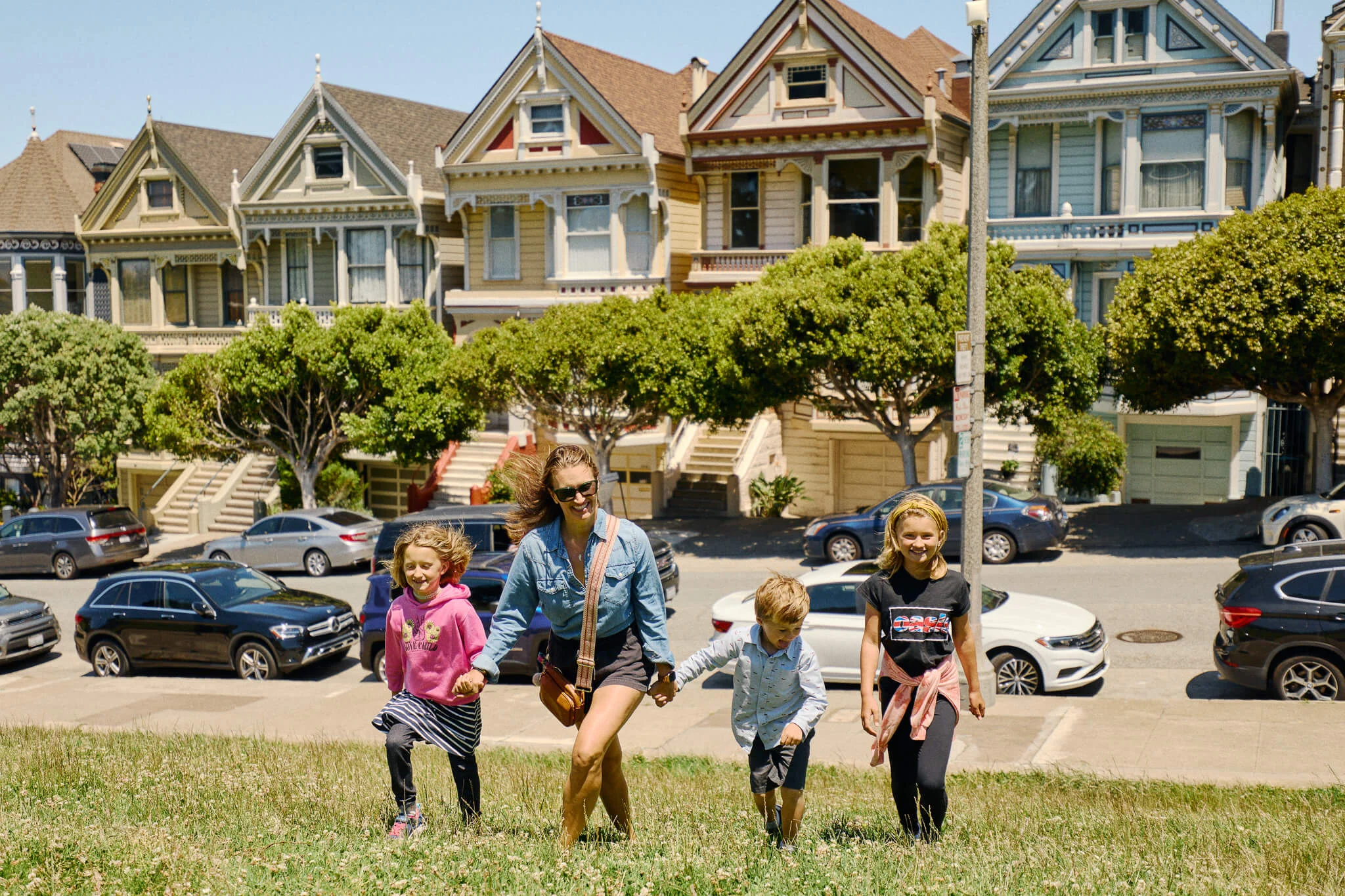 Woman and three children walk up a grassy hill holding hands in front of the colorful Victorian Painted Ladies houses, with parked cars and leafy trees lining the street behind them.