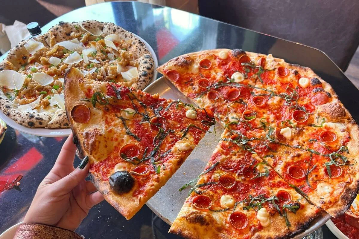 Overhead view of a person holding a large slice of thin-crust pepperoni pizza over a metal tray with the rest of the pie, with a second artisan pizza topped with shaved cheese and crumbled toppings on a plate in the background.