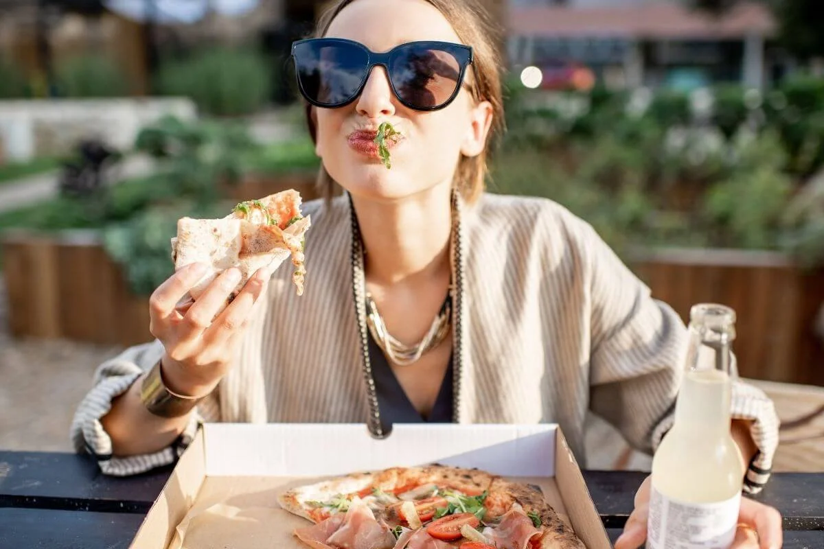 Young woman wearing large sunglasses sits at an outdoor table with an open pizza box, holding a slice of pizza in one hand and a bottled drink in the other, with herbs and sauce playfully stuck to her lips.