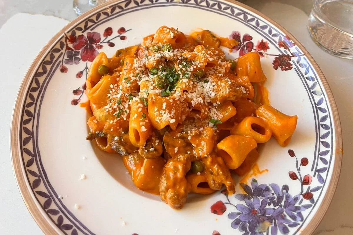 Close-up of a plate of rigatoni pasta coated in creamy tomato sauce with peas and mushrooms, topped with grated cheese and herbs, served on a white floral-patterned dish.