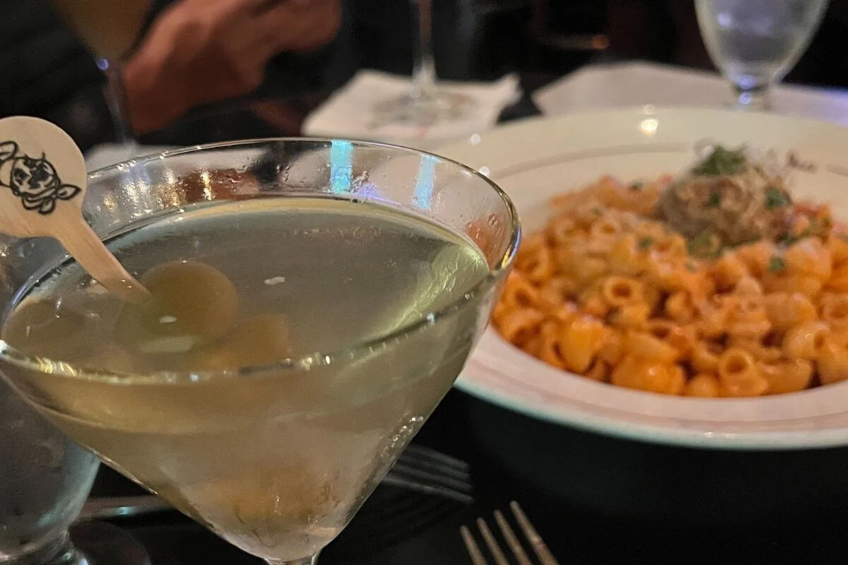 Close-up of a chilled martini with green olives in a cocktail glass, with a blurred plate of creamy tomato pasta in the background on a dimly lit restaurant table.