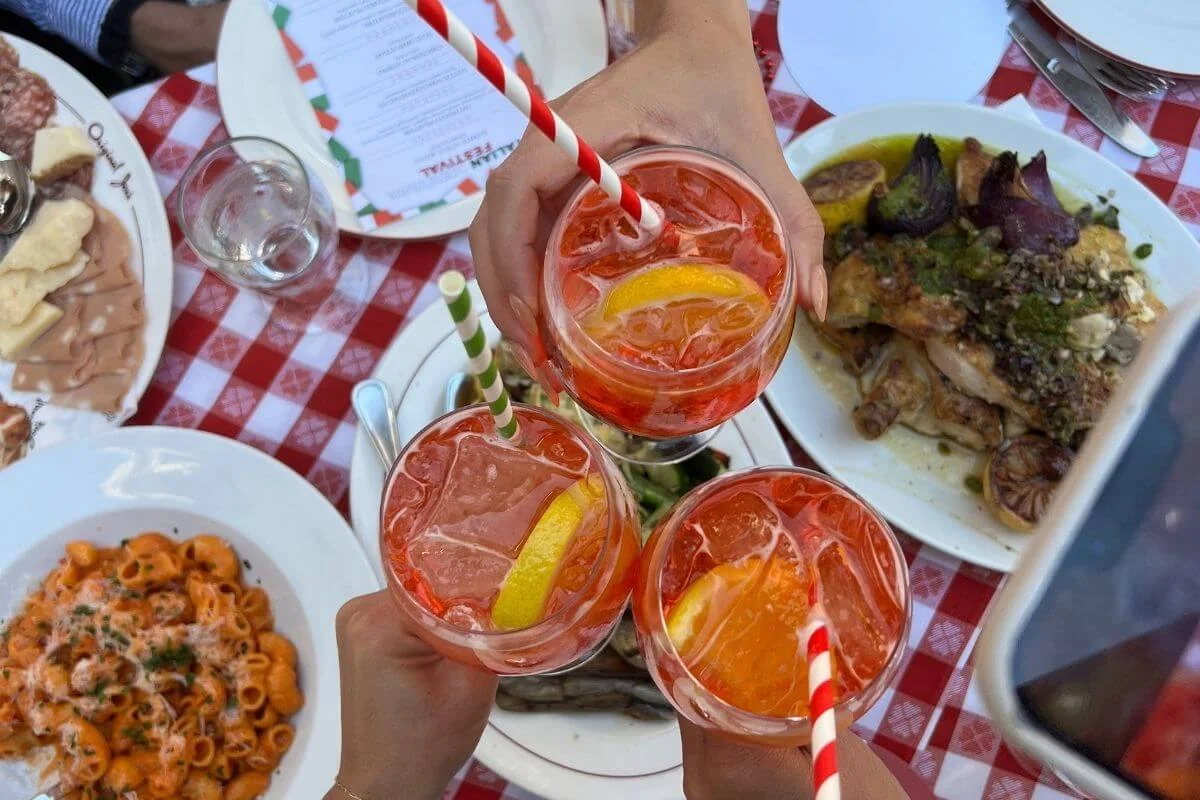 Overhead view of three people toasting with bright orange spritz cocktails with striped straws over a red-and-white checkered table set with pasta, roast chicken, and antipasto dishes.