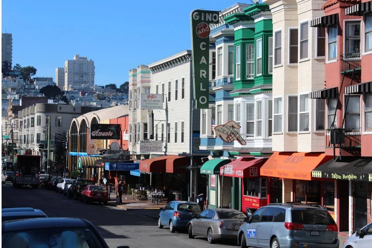 Daytime view of a busy street in San Francisco’s North Beach neighborhood, with colorful row buildings, restaurant awnings, classic bar signs, and parked cars lining the road against a hilly city backdrop.